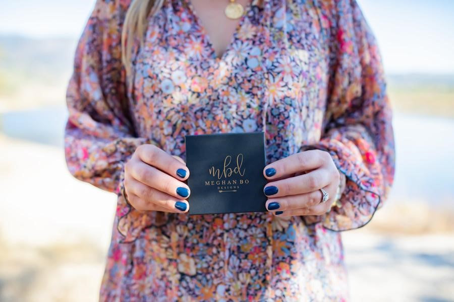 A woman wearing a long floral Free People dress holding a black Meghan Bo Designs jewelry box with blue dip nails. 