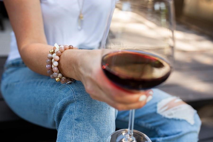 A woman wearing a white tee shirt and jeans sits with a glass of red wine and is wearing a stack of beaded stretch bracelets in pinks and whites made by Meghan Bo Designs.