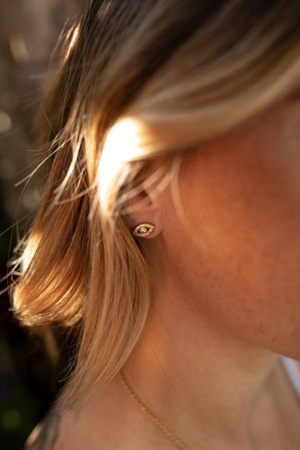 A woman with her brown hair pulled back is wearing Meghan Bo Designs evil eye cubic zirconia stud earrings in gold.