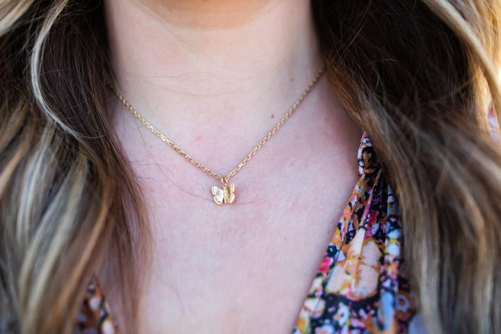 A woman with long wavy dirty blonde hair is wearing a floral Free People Dress with a gold necklace that has a small gold fill butterfly pendant attached called the Flutter Necklace by Meghan Bo Designs.
