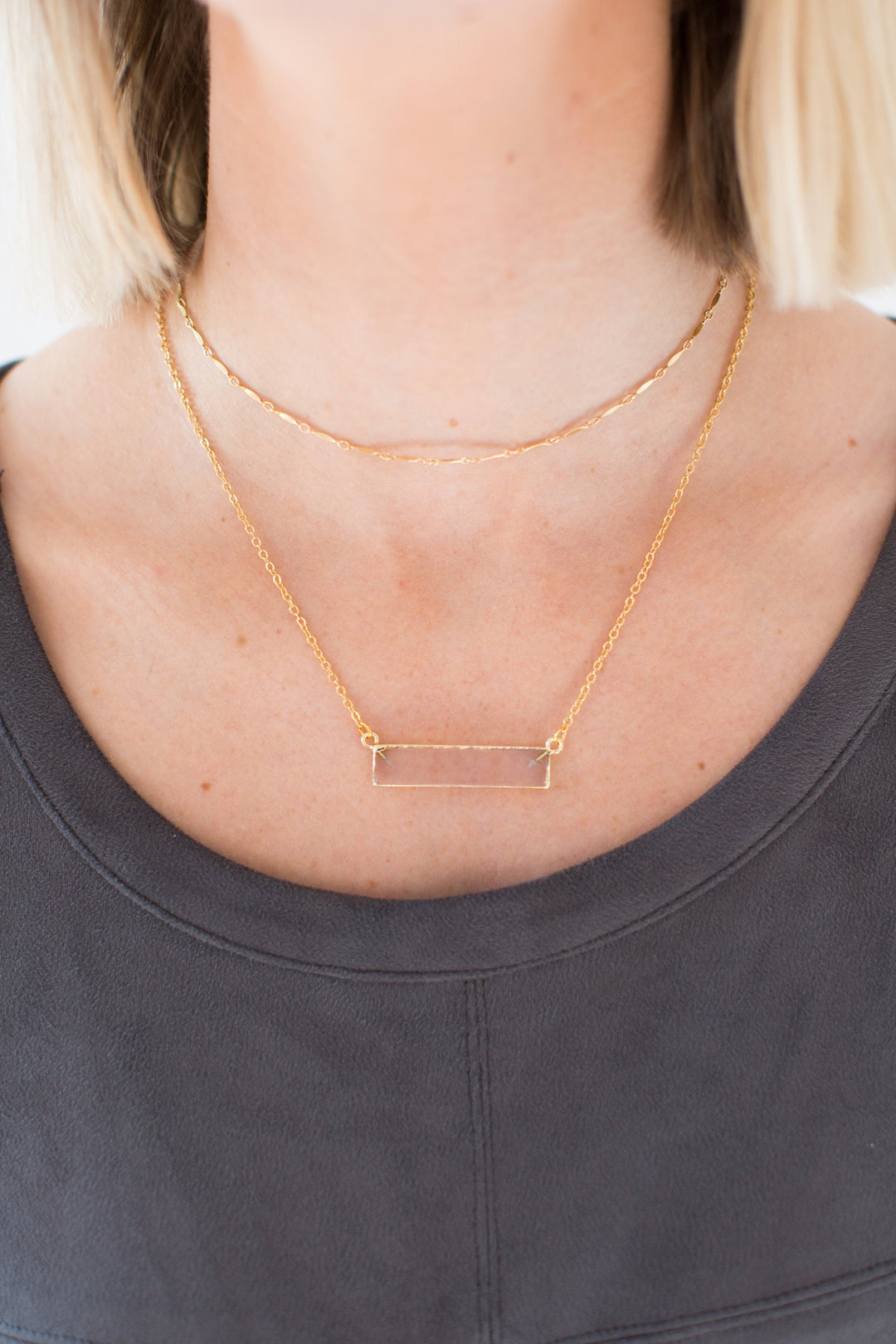 A woman with short blonde hair is wearing a gray dress by vici and two layered necklaces; one is a gold fill choker chain and the other is a clear quartz bar necklace made by Meghan Bo Designs. 