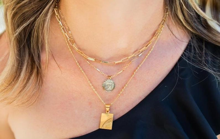 A woman with brown wavy hair is wearing an off the shoulder black shirt and three layered necklaces, a paperclip chain choker, a small gold coin necklace and a rectangle pendant necklace made by Meghan Bo Designs.