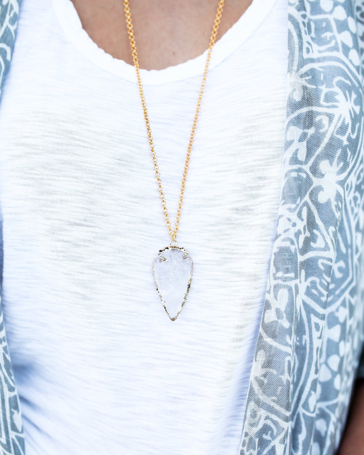 A woman is wearing a white scoop neck tee shirt from vici dolls with a long gold necklace that has a clear quartz arrowhead pendant edged in gold made by Meghan Bo Designs.