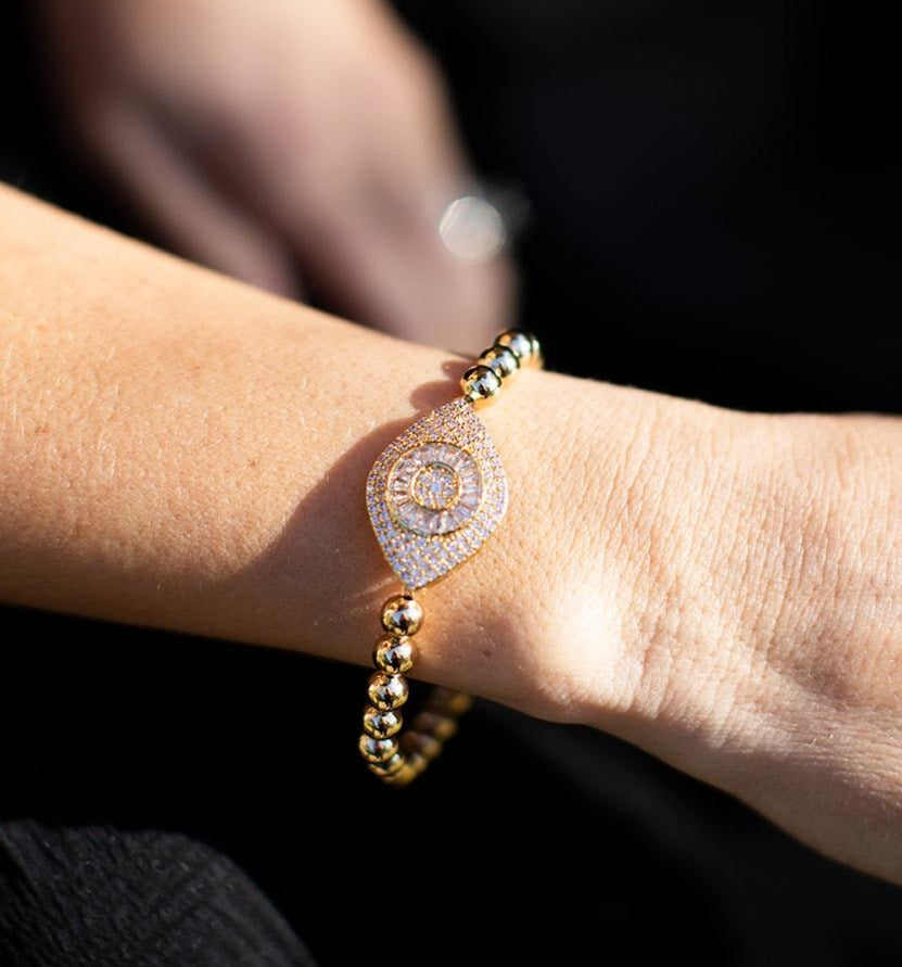A woman wearing a black VICI Collection dress sits with her right arm out wearing a gold beaded bracelet with a large cubic zirconia evil eye in the middle of it made by Meghan Bo Designs.