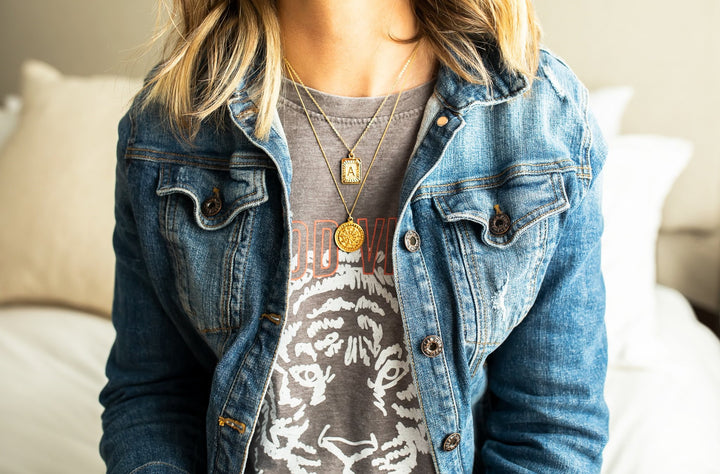 A woman with blonde hair wearing a VICI Collection denim jacket and a lulu and simon graphic tee shirt with two gold layered necklaces made by Meghan Bo Designs.