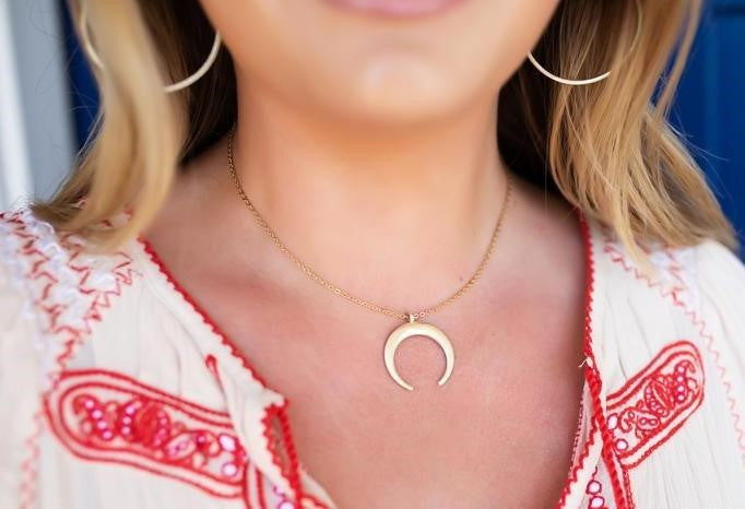 A woman wearing a white dress with red embroidery is wearing a pair of brushed gold hoop earrings and a short gold necklace with a gold crescent pendant made by Meghan Bo Designs.