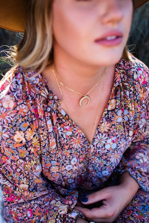 A woman with long blonde hair is wearing a brown hat, a floral Free People dress and two layered gold necklaces, the shorter necklace has a gold crescent moon pendant made by Meghan Bo Designs.