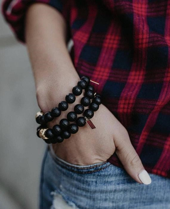 A woman wearing a red plaid shirt and jeans has her right hand in her pocket and is wearing a stack of bracelets with black beads and one gold bead made by Meghan Bo Designs.
