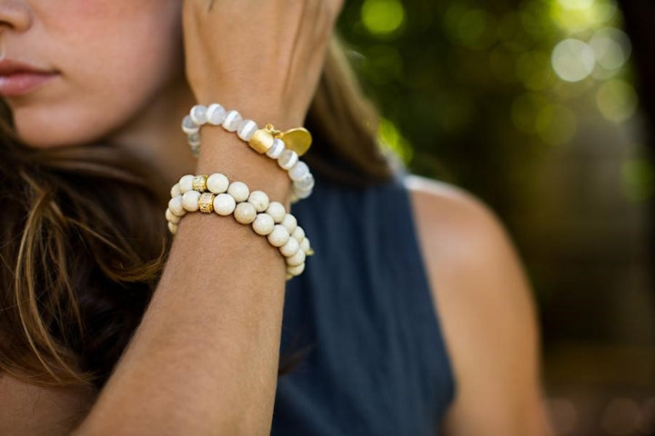 A woman with long brown hair brushes her hair back with her right hand and is wearing a stack of bracelets made with ivory colored fossil stone beads by Meghan Bo Designs.