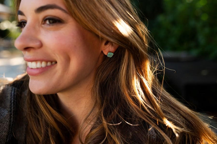 A woman with long brown hair looks to the side smiling, she is wearing a pair of round turquoise stud earrings that have a gold edge on them made by Meghan Bo Designs.