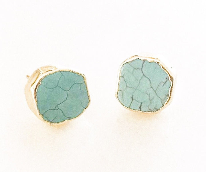 A pair of round turquoise stud earrings that have a gold edge lay on a white background made by Meghan Bo Designs.