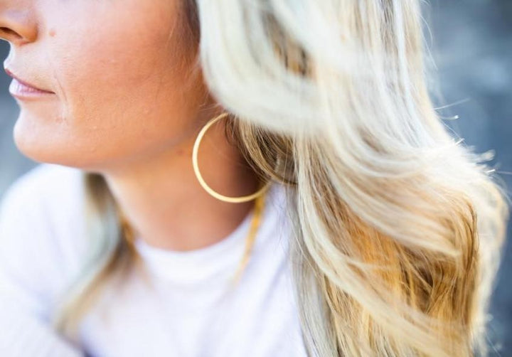 A blonde woman looks off to the side, she is wearing a gold necklace, white vici collection tee shirt and gold hoop earrings made by Meghan Bo Designs.