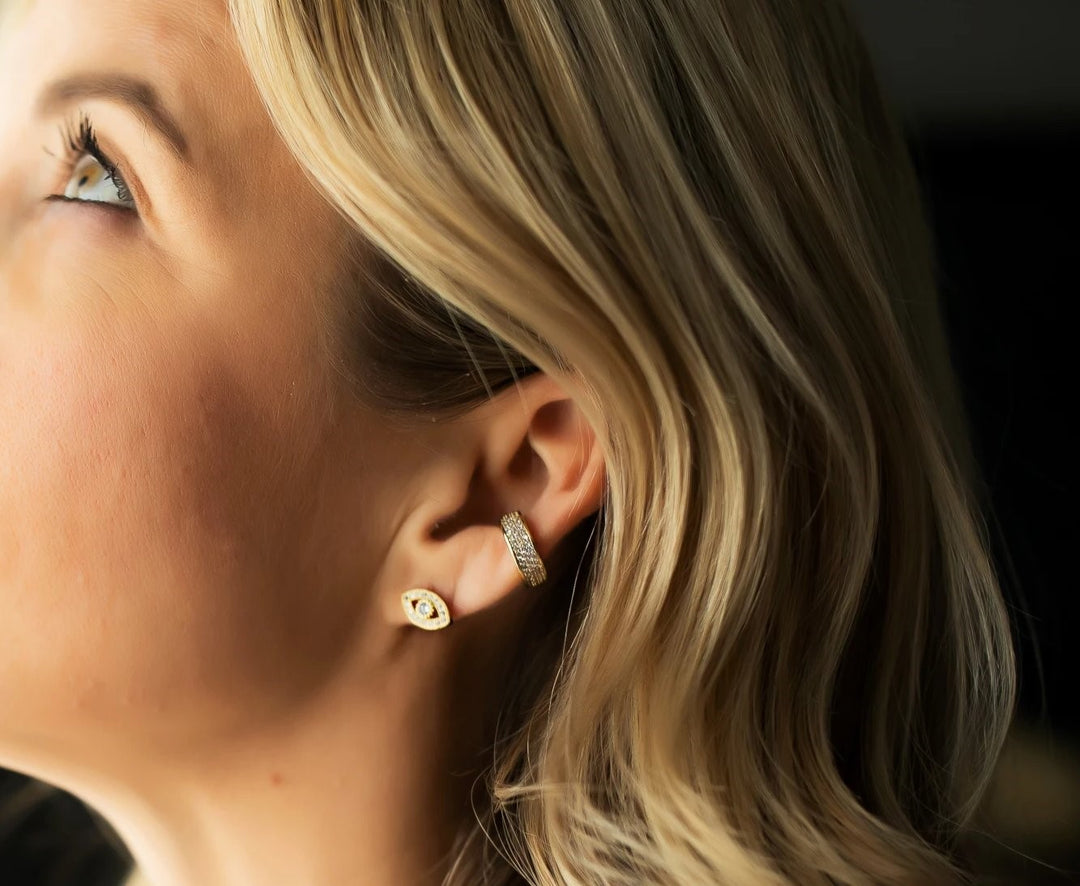 A woman with blonde wavy hair is looking up and to the side, she has a pair of cubic zirconia evil eye studs and a cz ear cuff made by Meghan Bo Designs.