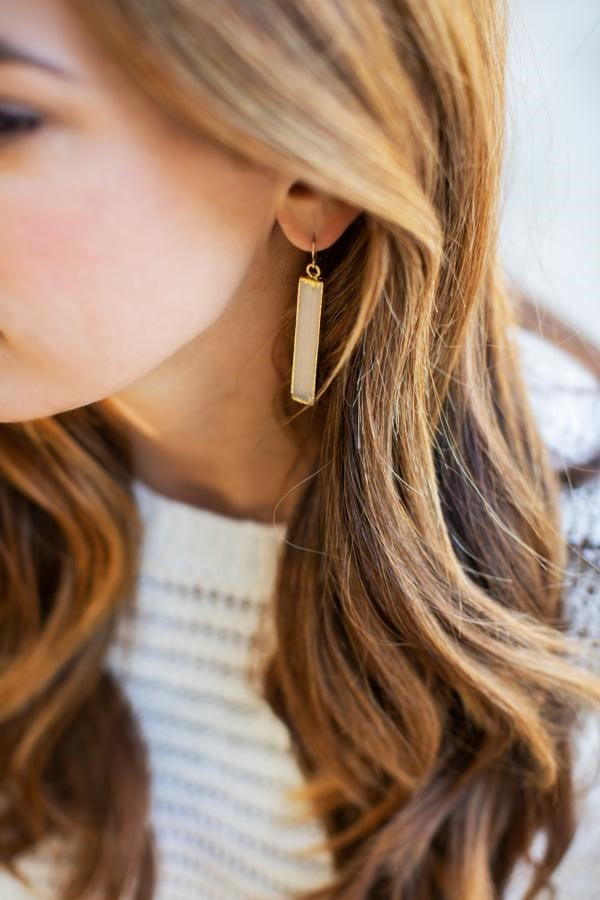 A woman with long brown wavy hair is wearing a white sweater and selenite crystal dangle earrings made by Meghan Bo Designs.