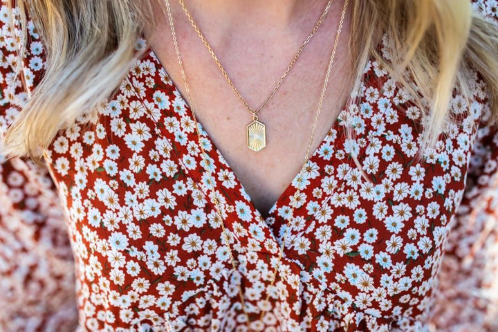 A woman with blond hair is wearing a floral vici collection dress with two gold necklaces, one has a shield pendant made by Meghan Bo Designs.