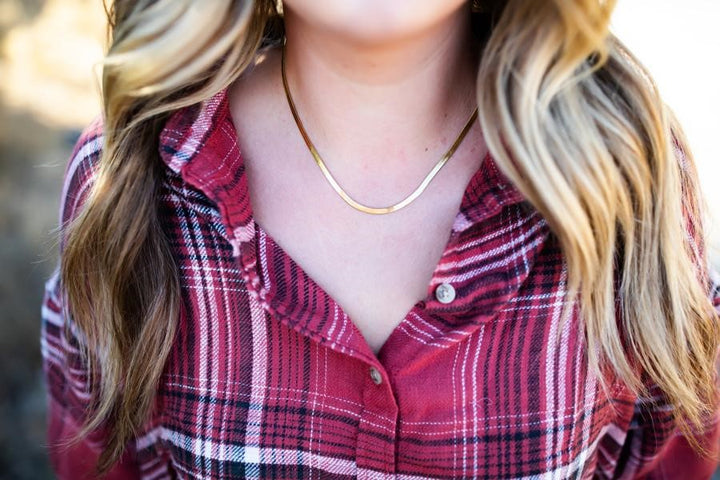 A woman with long blonde wavy hair is wearing a red plaid flannel shirt with a gold herringbone chain necklace made by Meghan Bo Designs.