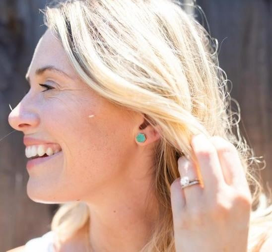 A woman with blonde hair is smiling and looking to the side, she has a solitaire diamond ring and turquoise stud earrings made by Meghan Bo Designs.